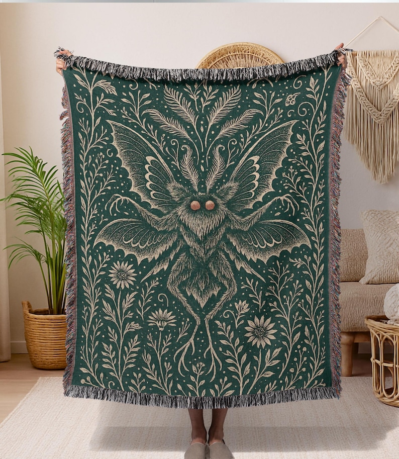 Puede incluir: Una manta tejida de color verde oscuro con un gran dise&ntilde;o de polilla y detalles florales y de hojas en beige claro. La manta tiene un borde con flecos y est&aacute; sostenida por una persona.