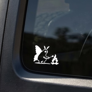 May include: A white vinyl decal featuring a moth-like creature with wings and antennae sitting by a small campfire. The creature is holding a stick with a marshmallow over the flames. The design is a solid white silhouette applied to a dark window.