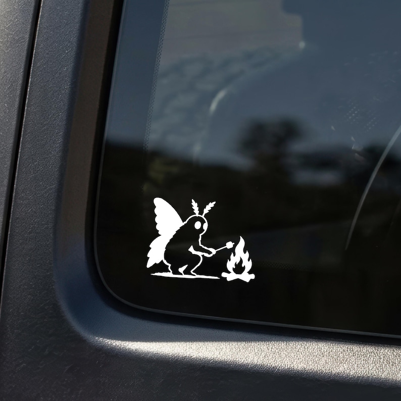 May include: A white vinyl decal featuring a moth-like creature with wings and antennae sitting by a small campfire. The creature is holding a stick with a marshmallow over the flames. The design is a solid white silhouette applied to a dark window.