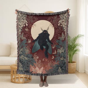 May include: A woven tapestry throw blanket featuring a dark silhouette of a moth-like creature with glowing eyes against a full moon. The design includes butterflies and foliage in shades of burgundy, teal, and gray, with a fringed edge.