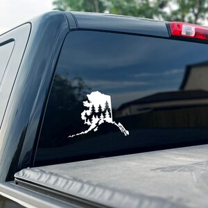 May include: A white vinyl decal applied to a dark vehicle window, featuring the silhouette of the state of Alaska filled with a detailed graphic of a dense evergreen forest with multiple pine trees layered within the state border shape.