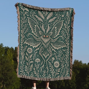 Puede incluir: Una manta verde oscuro con un dise&ntilde;o detallado de polilla y flores. La polilla tiene alas grandes y est&aacute; rodeada de elementos bot&aacute;nicos. La manta tiene un borde con flecos y se muestra contra un cielo azul.