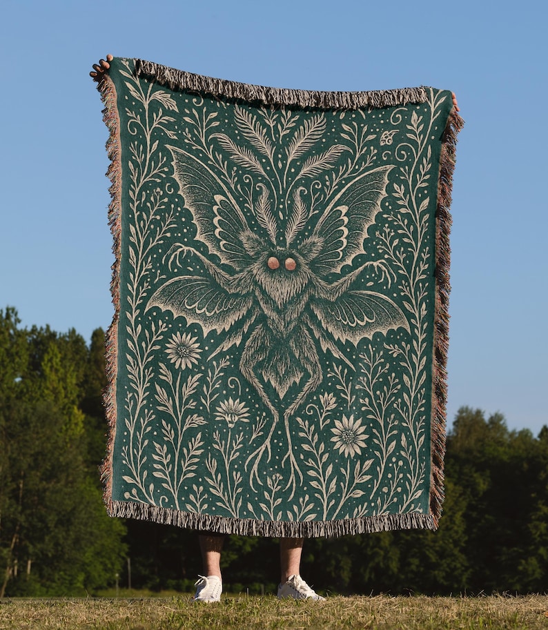 Puede incluir: Una manta verde oscuro con un dise&ntilde;o detallado de polilla y flores. La polilla tiene alas grandes y est&aacute; rodeada de elementos bot&aacute;nicos. La manta tiene un borde con flecos y se muestra contra un cielo azul.
