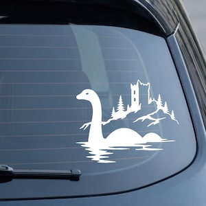May include: A white vinyl decal applied to a car window, featuring a silhouette of the Loch Ness monster emerging from water, with a castle tower and pine trees positioned on a rocky shoreline behind the creature's long neck and humped back.