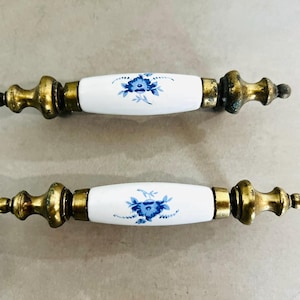 May include: Two antique cabinet pulls. Each pull features a white ceramic center with a blue floral design, flanked by ornate brass-toned metal ends. The pulls are designed for easy installation and add a touch of vintage charm to any furniture piece.