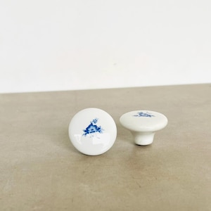 May include: Two white ceramic cabinet knobs with a blue floral design. One knob is round, and the other has a short stem. The knobs are on a neutral surface, perfect for adding a touch of elegance to furniture.