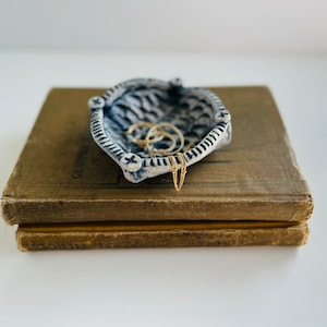 May include: A white ceramic dish with a blue and white geometric pattern, shaped like a boat, with gold rings inside. The dish is sitting on top of two brown books.