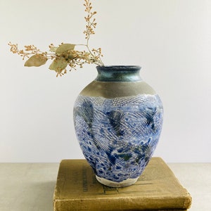 May include: A blue and white ceramic vase with a wavy pattern sits on top of two vintage books. A dried branch with small brown leaves is in the vase.