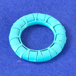 May include: A turquoise-colored, circular object with a woven or segmented design. The object has a hollow center and appears to be made of a solid material. The background is a solid blue color. The text "COPREERKEY.COM" is visible at the bottom.