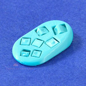 May include: A turquoise, oval-shaped 3D-printed item featuring square and cube-shaped recesses. The object is set against a blue background. The surface of the item has a textured, ridged pattern.