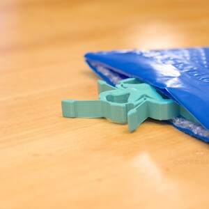 Pu&ograve; includere: Un oggetto a forma di stella, di colore verde acqua chiaro, emerge parzialmente da una busta a bolle blu. L'oggetto &egrave; fatto di un materiale solido e ha un design geometrico. La busta poggia su una superficie di legno marrone chiaro.