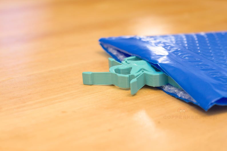 Pu&ograve; includere: Un oggetto a forma di stella, di colore verde acqua chiaro, emerge parzialmente da una busta a bolle blu. L'oggetto &egrave; fatto di un materiale solido e ha un design geometrico. La busta poggia su una superficie di legno marrone chiaro.