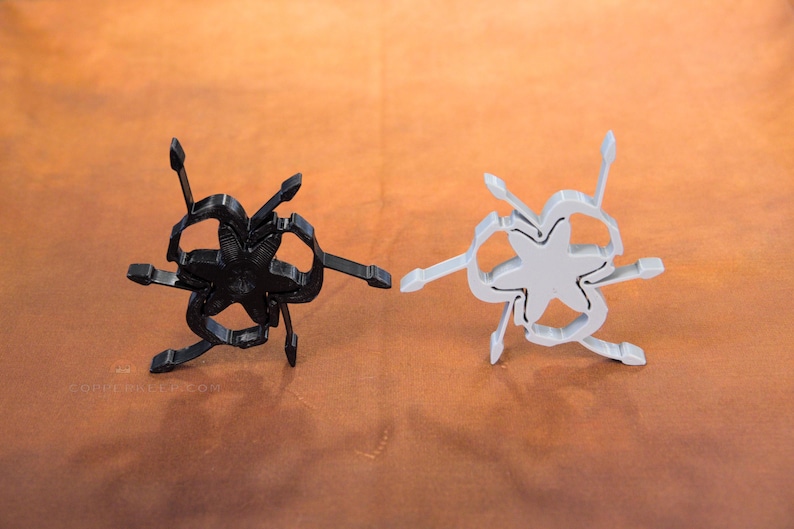 Pu&ograve; includere: Due oggetti astratti a forma di stella. Uno &egrave; nero, l'altro grigio chiaro. Ognuno ha pi&ugrave; braccia che si estendono verso l'esterno. Gli oggetti sono esposti su una superficie marrone. Il design &egrave; moderno e geometrico.