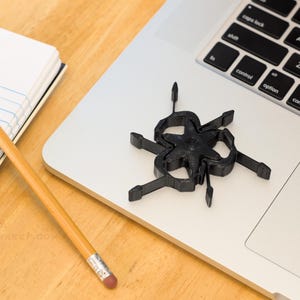 Pu&ograve; includere: Un oggetto nero a forma di stella con pi&ugrave; bracci &egrave; appoggiato su un laptop. Sono visibili anche una matita gialla con una gomma rosa e un blocco note con carta a righe blu. Gli oggetti sono su una superficie di legno di colore chiaro.