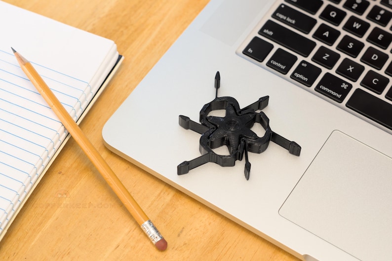 Pu&ograve; includere: Un oggetto nero a forma di stella con pi&ugrave; bracci &egrave; appoggiato su un laptop. Sono visibili anche una matita gialla con una gomma rosa e un blocco note con carta a righe blu. Gli oggetti sono su una superficie di legno di colore chiaro.