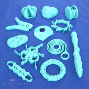 May include: A collection of various turquoise 3D-printed objects, including a flower-shaped item, a ring, a wand, and other geometric shapes. The objects are arranged on a blue surface.