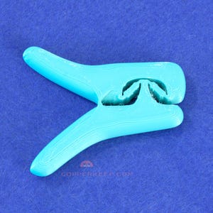 May include: A turquoise plastic clothes peg with a distinctive design. The peg has a curved shape with a central opening and two extended arms. The surface shows layer lines from 3D printing. The text "COPPERKEEP.COM" is visible on the bottom.
