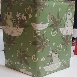 May include: A green fabric notebook cover with a repeating pattern of frogs smoking marijuana leaves in bowls, hearts, and marijuana leaves.