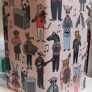May include: A pink fabric book cover with a whimsical design featuring various animals playing musical instruments. The animals include a badger, a cat, a deer, a zebra, a rabbit, a panda, an owl, an elephant, and a bird. The fabric has a light pink background with musical notes and a repeating pattern of the animals.