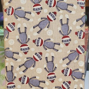 May include: A beige fabric item covered in a repeating pattern of sock monkey illustrations. The sock monkeys are gray with red and white hats and mouths. White buttons are scattered throughout the design.
