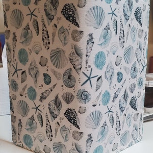 May include: A white fabric notebook cover with a blue and gray seashell pattern. The cover features a variety of seashells, including clams, scallops, and starfish.