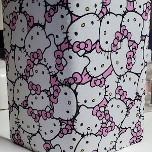 May include: A white fabric pouch with a repeating pattern of Hello Kitty faces in pink and black. The pouch is made of a soft, textured fabric.