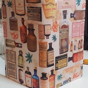 May include: A vintage fabric design featuring a repeating pattern of antique medicine bottles with labels that read "Cannabis Indica", "Cannabis Sativa", "Doctor Bagilister's Cough Mixture", and "Cannabis Americana". The fabric is a light beige color with a subtle orange and brown tie-dye effect.