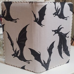 May include: A beige fabric journal cover with a black stitched border. The cover features a repeating pattern of black bats with outstretched wings.