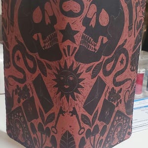 May include: A red leather notebook cover with a black and white design featuring two skulls, a sun, keys, and other symbols. The cover has a stitched edge.