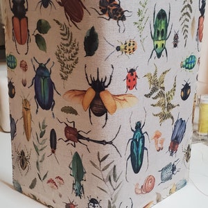 May include: A beige linen fabric notebook cover with a repeating pattern of colorful insects, including beetles, butterflies, and ladybugs. The cover has a stitched edge and a subtle texture.