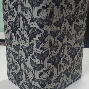 May include: A square, gray container with a repeating pattern of black butterflies and ink splatter designs. The butterflies vary in size and are detailed with intricate wing patterns. The container has a textured appearance.