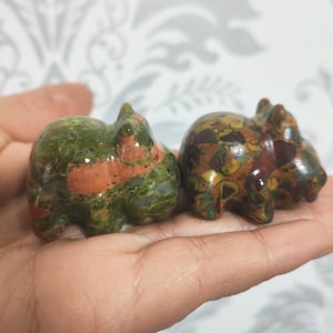 以下が含まれることがあります： 緑、茶色、赤のジャスパーで精巧に彫刻された、小さなカバの置物2つ。置物は手のひらの上に置かれています。