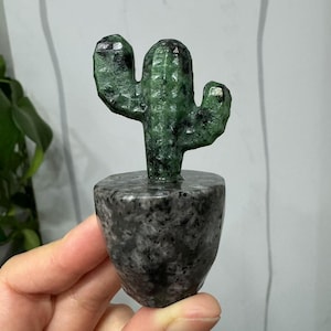May include: A green gemstone cactus sculpture with a black and grey speckled base. The cactus is carved in a realistic style with multiple arms.