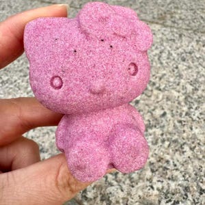 May include: A pink Hello Kitty bath bomb shaped like a cartoon character. The bath bomb has a textured surface and is made of a soft, powdery material.
