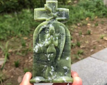 May include: A green stone sculpture of an angel with outstretched wings, praying. The angel is set within an arched frame, topped with a cross and a dove. The sculpture is held in a hand, with a blurred green background.