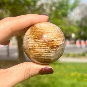 May include: A sphere of banded agate with brown and white stripes. The sphere is smooth and polished, and it is being held in a hand.