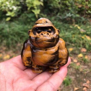 Könnte beinhalten: Eine kleine, handgeschnitzte Gorillafigur aus poliertem Tigerauge-Edelstein. Die Skulptur weist die charakteristischen goldbraunen und schwarzen schimmernden Bänder des Steins auf und ist als sitzender Gorilla mit rundem Kopf geformt.