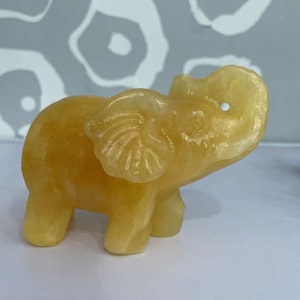 Op de afbeelding: Een klein, geel, gesneden olifantenbeeldje. De olifant staat met zijn slurf omhoog.