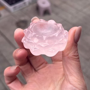 Op de afbeelding: Een lichtroze, gebeeldhouwde stenen sculptuur van een lotusbloem. De sculptuur wordt in een hand gehouden en toont de ingewikkelde details van de bloemblaadjes en de centrale verdieping. De steen heeft een doorschijnende kwaliteit.