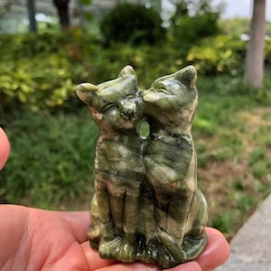 Può includere: Una scultura in pietra scolpita di due gatti che si baciano. I gatti sono di colore verde screziato con venature verdi più scure. La scultura è tenuta in una mano, con uno sfondo verde sfocato.