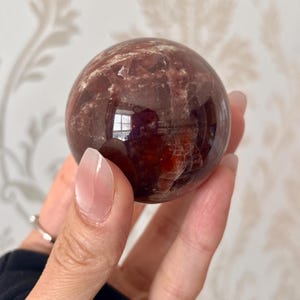 May include: A polished, spherical gemstone with a deep reddish-brown hue, featuring internal patterns and a reflective surface. The sphere is held in a hand, showcasing its size and clarity. This crystal ball is a decorative item.