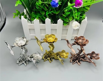 May include: Three decorative metal stands in silver, gold, and copper finishes. Each stand features a stylised tree branch with a bird perched on each branch and a flower on top. The stands are approximately 10 cm tall.