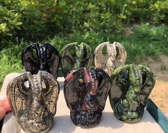 May include: A collection of carved stone dragon figurines in various colors, including black, green, and tan. The dragons have detailed wings and are displayed on a green surface against a backdrop of greenery.