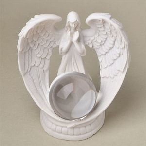 May include: White porcelain angel figurine with outstretched wings holding a clear glass orb. The angel is kneeling with hands clasped in prayer.