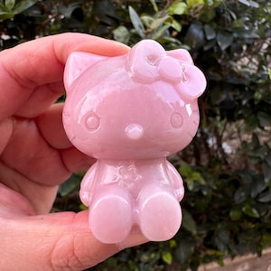 Puede incluir: Una figurita rosa de Hello Kitty hecha de piedra pulida. La figurita está sentada en una mano y tiene un lazo en la cabeza.