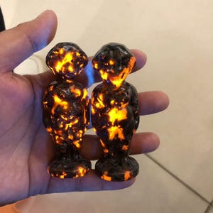 May include: Two small, dark sculptures with a glowing orange and yellow interior. The figures have a rounded head and body, standing on a circular base. The sculptures are held in a hand, with the light reflecting off the surface.