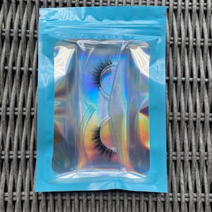 May include: Two pairs of false eyelashes in a clear plastic pack with a holographic iridescent background. The pack is inside a light blue resealable plastic bag.