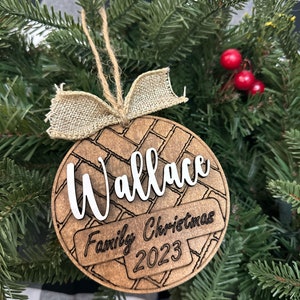 Puede incluir: Un adorno navideño de madera con un patrón de ladrillo marrón y blanco. El adorno está personalizado con el nombre "Wallace" y el texto "Navidad en familia 2023".
