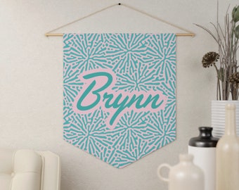 May include: A wall hanging banner with a pink and teal pattern and the name "Brynn" in teal script. The banner is suspended from a wooden rod with a natural cord.