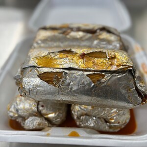 May include: A stack of tamales wrapped in silver foil, presented in a white foam container. The tamales have a golden-brown filling visible through the foil. The container has a small amount of sauce at the bottom.